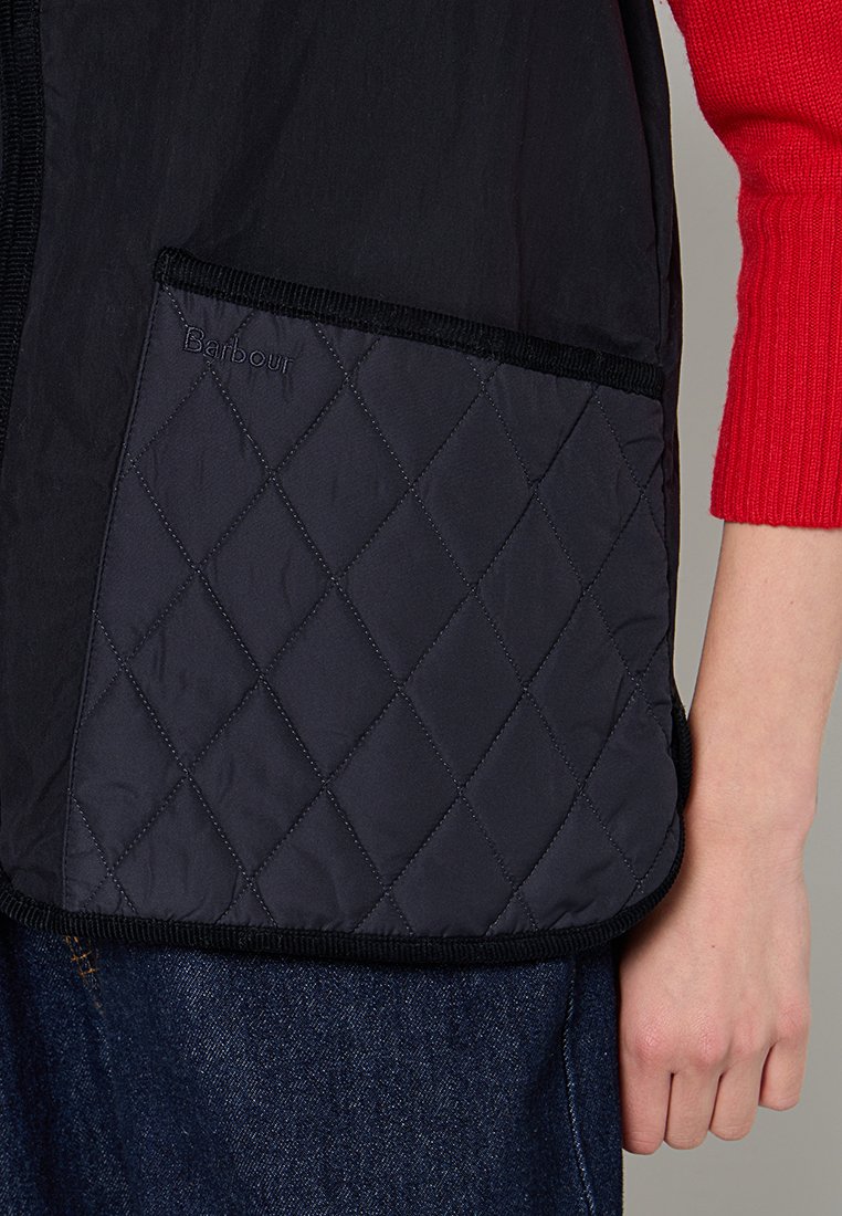 Czarna, pikowana kamizelka z granatową kieszenią w romby i haftowanym logo "Barbour", wykończona czarną lamówką. Widoczny rękaw czerwonego swetra.