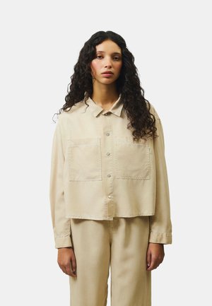 Donna con capelli lunghi e ricci che indossa una camicia beige con bottoni e grandi tasche anteriori e pantaloni beige abbinati, in piedi su uno sfondo bianco.