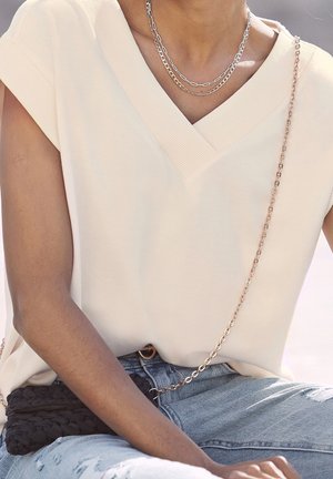 Persona que lleva una blusa sin mangas de color crema con cuello en V, collares de cadena plateados en capas, vaqueros azules rasgados y un bolso tejido negro con una cadena dorada como asa.