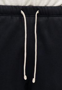 Μαύρο παντελόνι φόρμας με ελαστική μέση και μακριά λευκά κορδόνια που κρέμονται στο μπροστινό μέρος.