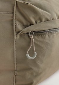 Geanta din țesătură khaki cu un buzunar cu fermoar. Cotelează un cârlig gri și cusături de-a lungul cusăturilor. Textură netedă cu un aspect ușor șifonat.