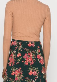 Top lungo a maniche ribbed marrone abbinato a una gonna floreale verde. La gonna presenta fiori rossi e rosa su uno sfondo verde scuro.