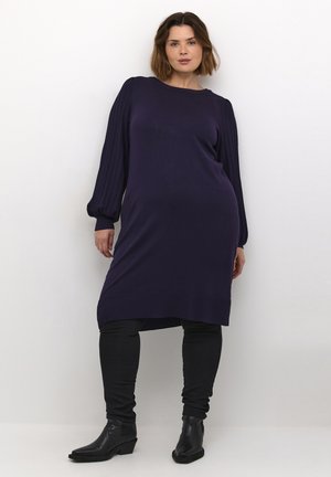 Langärmliges, dunkel-lila Strickkleid mit gerippten Ärmeln, rundem Ausschnitt und seitlichen Schlitzen. Kombiniert mit schwarzen Skinny-Jeans und schwarzen Stiefeletten.