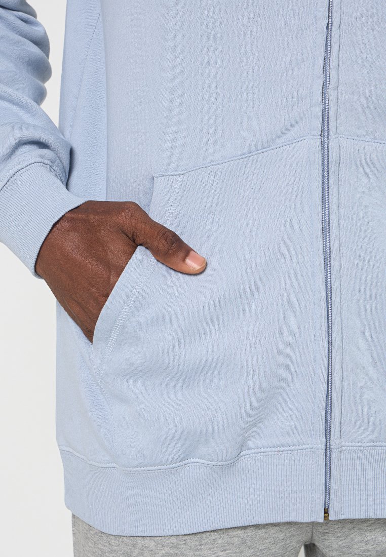 Hellblaues Zip-Hoodie mit weicher Textur, Kängurutasche und gerippten Bündchen. Nahaufnahme zeigt Details der Tasche und des Reißverschlusses.