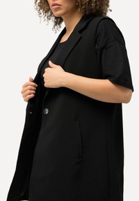 Svart skräddarsydd väst med strukturerad axeldetalj, knappstängning framtill, sidofickor och mjuk tygstruktur.