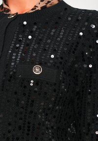 Cárdigan negro con lentejuelas y botón floral dorado sobre una blusa de leopardo, mostrado desde el hombro hasta el pecho.