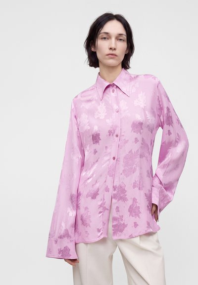 Femme aux cheveux courts et foncés portant un chemisier en satin rose clair à motif floral avec de larges poignets, associé à un pantalon couleur crème.