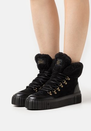 Zapatillas altas negras con puños de piel de peluche, materiales de ante y cuero, ojales en tono dorado y una suela de plataforma texturizada.