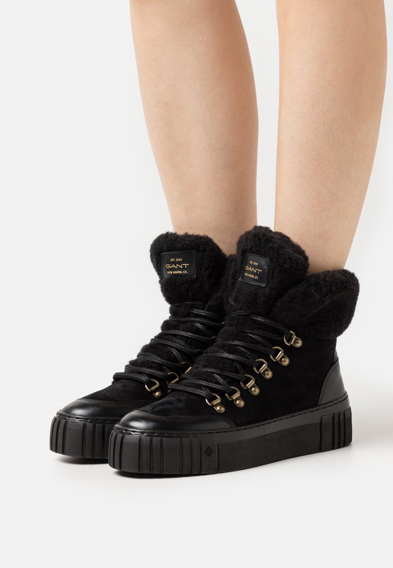 Zapatillas altas negras con puños de piel de peluche, materiales de ante y cuero, ojales en tono dorado y una suela de plataforma texturizada.
