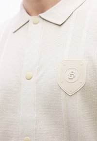 Κοντινό πλάνο ενό ς μπεζ πουκάμισου με υφή πλέξης, με κουμπιά και ένα σχέδιο σε σχήμα ασπίδας που φέρει ένα στυλιζαρισμένο γράμμα "B" και τέσσερα αστέρια στο στήθος.