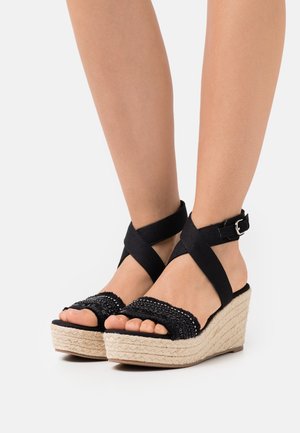 Sandales compensées en espadrilles noires avec des brides tressées, une boucle à la cheville et une plateforme en jute texturé. Le design à bout ouvert met en valeur les pieds nus.