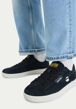 Schwarze G-Star-Sneaker mit weißen Sohlen, getragen mit weißen Socken und hellblauen, aufgerollten Jeans, auf weißem Hintergrund.