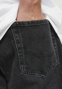 Chaqueta de mezclilla negra con un bolsillo trasero cuadrado y detalles de doble costura. La tela tiene una textura sutil y parece ligeramente desgastada.