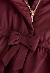 Burgunderfarbige Jacke mit einer strukturierten, glatten Oberfläche, die mit einer Schleifenverzierung an der Taille und einem goldenen Reißverschluss am Ausschnitt ausgestattet ist.