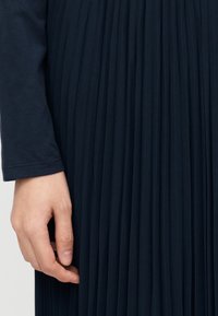 Ύφασμα φόρεμα σε σκούρο μπλε με πτυχώσεις που δείχνει ομαλή υφή με κάθετες πτυχώσεις· στο αριστερό μέρος φαίνεται μερικώς ένα χέρι που κρατάει το τελείωμα.