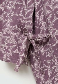 Fialová látka s květinovým vzorem v bílé, zdobená uzlem na okraji. Materiál vypadá texturovaně a je jemný.