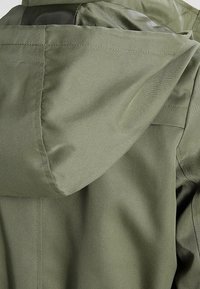 Veste à capuche vert olive avec une texture lisse et mate, coutures visibles et coupe ajustée au niveau du cou et des épaules.