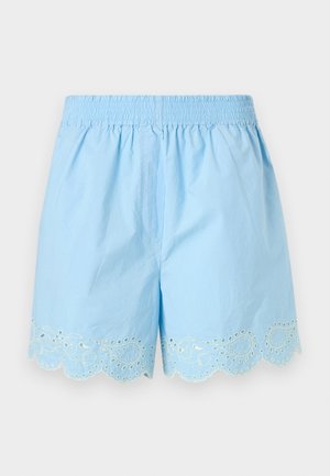 Modelo con camisa azul claro de botones y cuello con borde festoneado, y pantalones cortos a juego con ribete de encaje ojales, combinados con zapatillas deportivas beige.