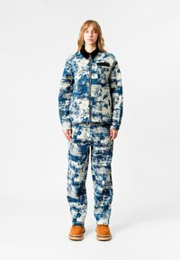 Chaqueta y pantalones de patrón azul y blanco, hechos de un tejido texturizado, con cierre frontal de cremallera y cuello negro. Presenta botas color tostado.