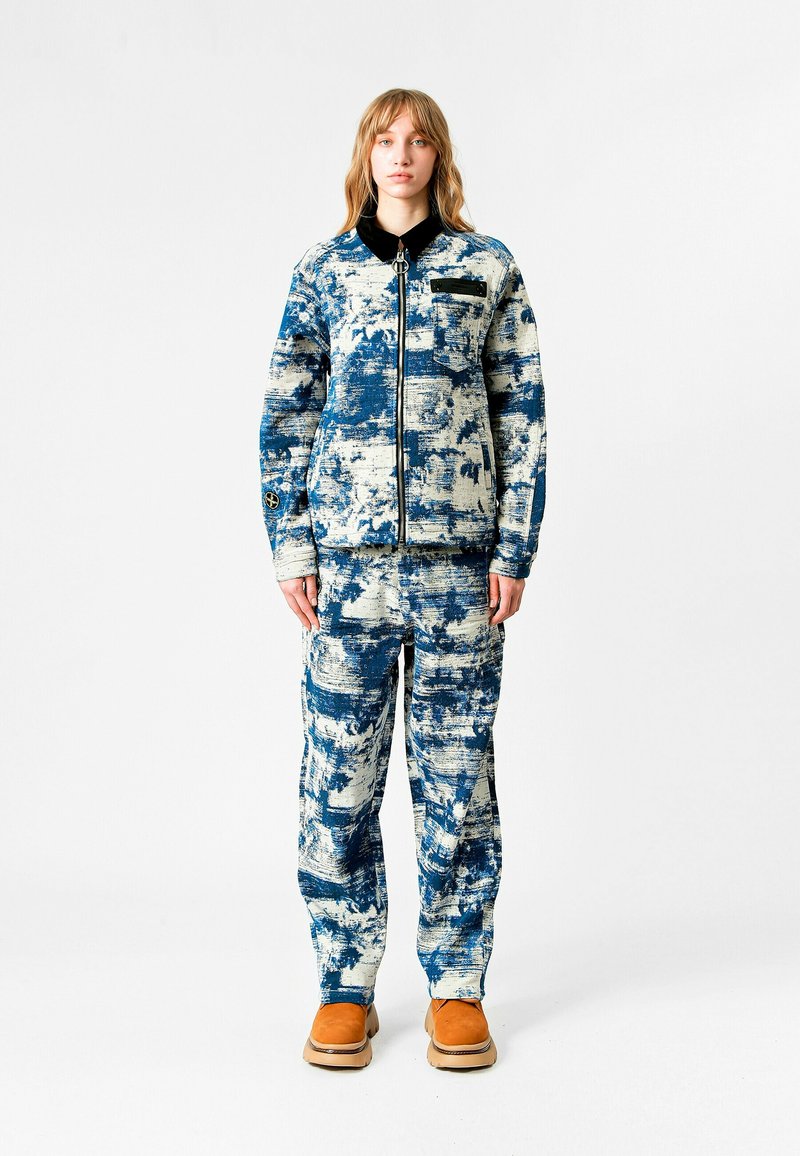 Chaqueta y pantalones de patrón azul y blanco, hechos de un tejido texturizado, con cierre frontal de cremallera y cuello negro. Presenta botas color tostado.
