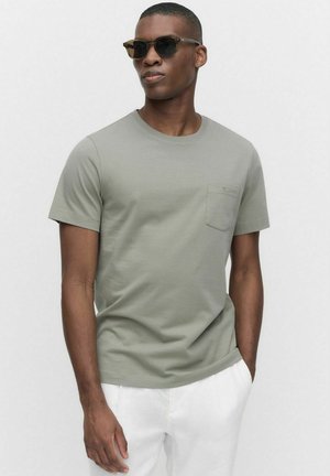 Lys grøn bomulds t-shirt med rund halsudskæring, korte ærmer og en venstre brystlomme. Modellen bærer sorte solbriller og hvide bukser.