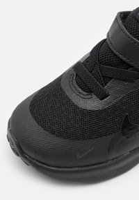Chaussure de sport noire en matériau en mesh, surface texturée, embout renforcé et sangle Velcro. Comprend un détail de logo sur le côté.