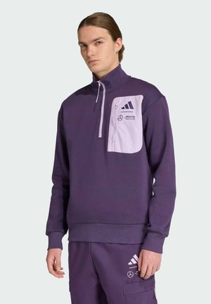 Fioletowy sweatshirt z wysokim kołnierzem, zamkiem błyskawicznym do jednej czwartej i białą kieszenią na piersi z logo. Noszony z dopasowanymi fioletowymi spodniami.