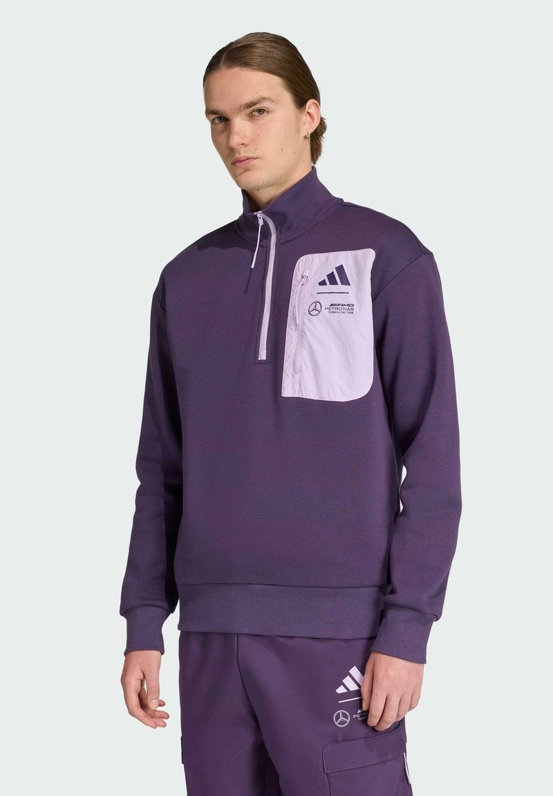 Felpa viola con colletto alto, zip a un quarto e taschino bianco sul petto con loghi. Indossata con pantaloni viola coordinati.