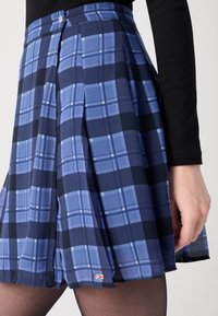 Plaid skirt in shades of blue and navy, featuring pleats, a fitted waist, and a small logo tag on the hem. Smooth fabric texture.
