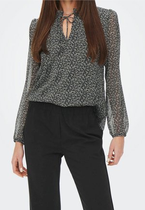 Vrouw draagt een zwarte bloemenblouse met lange mouwen en een striksluiting bij de hals en zwarte, hooggetailleerde broek, staand met ontspannen armen langs het lichaam.