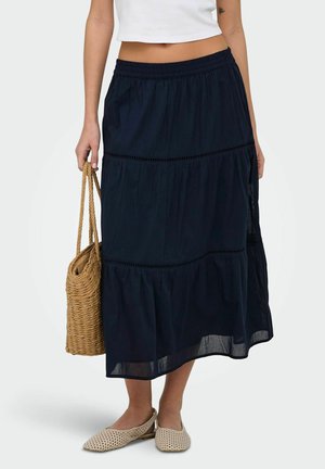 Femme portant une jupe midi à volants bleu marine, un débardeur blanc court, des chaussures plates tissées beige, et tenant un sac cabas tissé marron.