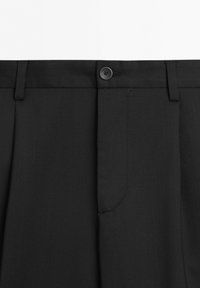 Close-up of black pants waistband with belt loops, a button fastener, and a hidden zipper fly on plain white background.