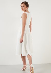 Rochie albă în stil A, cu un corset ajustat, design fără mâneci și lungime până la jumătatea gambei. Material neted cu un luciu subtil, asortată cu sandale cu curele.