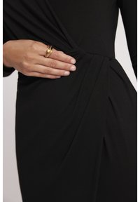 Czarna sukienka o drapowanej konstrukcji, teksturowanym materiale oraz detalem w postaci skrętu w talii. Na tkaninie spoczywa dłoń ozdobiona złotym pierścionkiem.