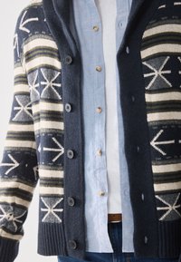 Wollcardigan mit Schalkragen, in marineblau und cremefarben, geometrischen Mustern und sechs schwarzen Knöpfen. Über einem hellblauen Hemd getragen.