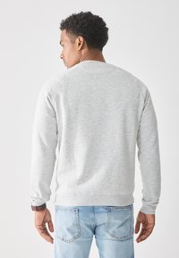 Hombre con pelo rizado corto, vistiendo un sudadera gris claro y jeans azul claro, de pie de espaldas contra un fondo blanco.