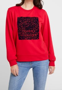 Rode sweatshirt met een grafisch ontwerp van een donker vierkant met een bloemmotief en de tekst "TOMMY HILFIGER" in het midden. Ribgebreide mouwen.
