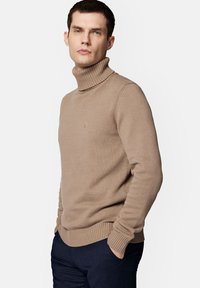 Beżowy sweter z golfem, z prążkowanymi mankietami i kołnierzem, wykonany z teksturowanej dzianiny. Dyskretny akcent logo na klatce piersiowej.