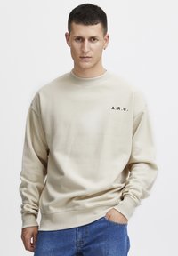 Sudadera beige con cuello redondo acanalado y mangas largas; presenta un logo negro impreso "A.R.C." en el lado izquierdo del pecho. Combinada con jeans azules.