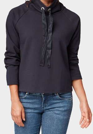 Osoba ubrana w ciemnogranatowy sweter z długim rękawem, z wysokim kołnierzem i szerokimi czarnymi trokami, w połączeniu z niebieskimi dżinsami.