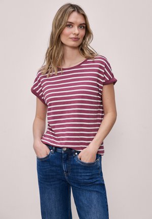 Femme aux cheveux blonds portant un t-shirt à manches courtes rayé bordeaux et blanc, et un jean bleu, debout les mains dans les poches.