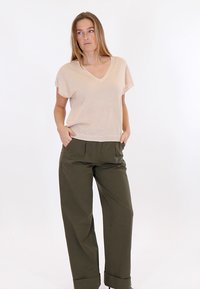 Kvinde står med hænderne i lommerne iført en beige kortærmet V-hals top og løse olivengrønne bukser mod en ensfarvet hvid baggrund.