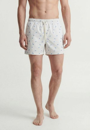 Man draagt witte zwembroek met blauwe schelpenpatronen, staat blootsvoets tegen een effen lichte achtergrond.