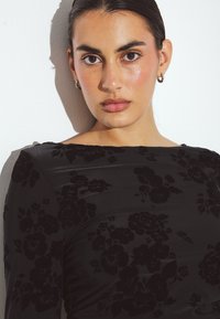 Črna obleka s cvetličnim vzorcem in mehkim teksturo. Ima dolge rokave in čolnasti izrez. Model prikazuje naravno ličenje in obroče v ušesih.