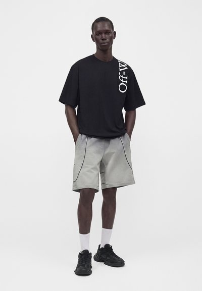 Άνδρας που φοράει oversized μαύρη μπλούζα με λευκό κείμενο Off-White, ανοιχτό γκρι σορτς με μαύρη λεπτομέρεια, λευκές κάλτσες και μαύρα αθλητικά παπούτσια, στέκεται με τα χέρια στις τσέπες.
