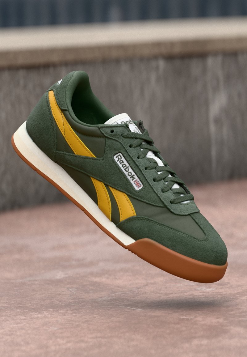 Sapatilhas verdes em camurça com detalhes amarelos e atacadores brancos. Apresenta uma sola de borracha castanha e o logótipo da Reebok. Design desportivo clássico.