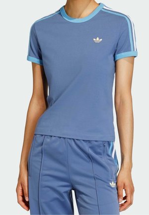 Personne portant un t-shirt Adidas bleu à manches courtes et un pantalon de survêtement assorti avec des rayures et logos bleu clair.