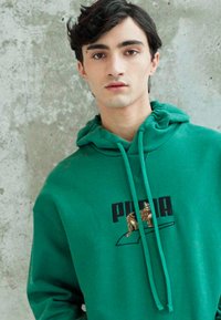 Felpa verde in cotone con tasca a marsupio, caratterizzata da un logo "PUMA" nero e una grafica di un leopardo. Il cappuccio è dotato di cordino e le maniche hanno polsini a costine.