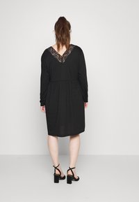 Vestido preto até ao joelho com mangas longas, detalhe em renda em V, cintura elástica e textura suave. Combinado com sandálias pretas de salto bloco.