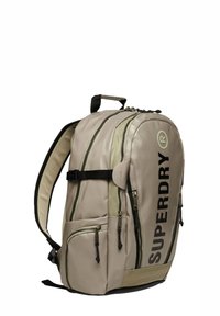 Zaino beige con accenti scuri; presenta più scomparti con cerniera, spallacci imbottiti e una maniglia superiore. Logo "SUPERDRY" ben visibile.